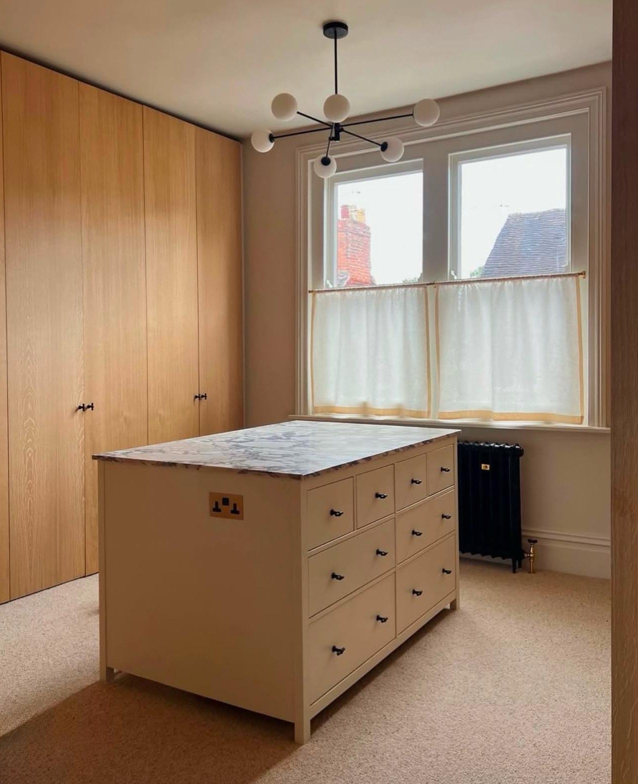 Beige dressing island with marble countertop in a dressing room with wooden wardrobes and large windows with cafe curtains.